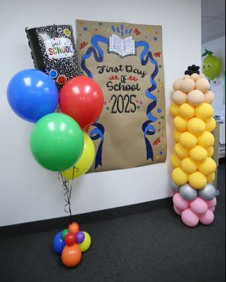 Colorful balloons and a decorated bag on a table with a 'First Day of School' sign.